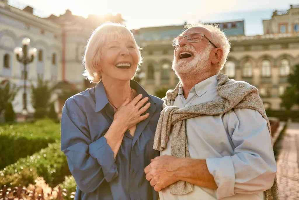 Beautiful and stylish senior couple laughing while standing in the park together on a sunny day, symbolizing the joys and freedoms of effective hearing loss treatment